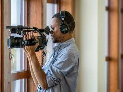 Student from School of Communication and Media wear headphones and holding a video camera filming through a window.